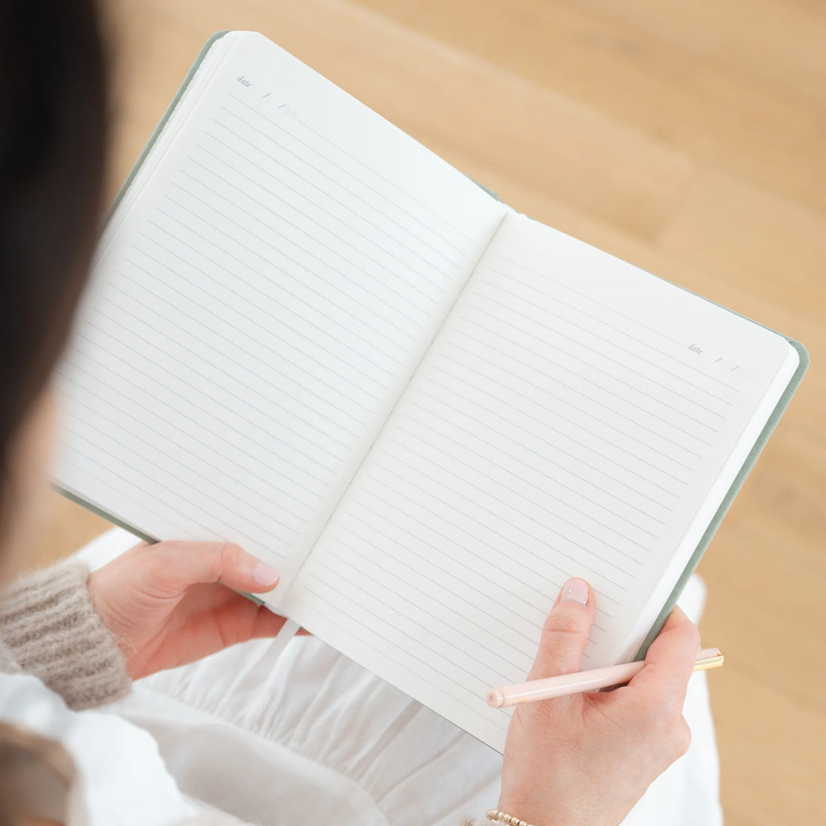 A person holding a pencil and an open Sugar Paper Sage Tailored Journal, a blank hardcover notebook with lined pages, viewed from above.