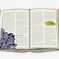 An open book, "The 100 Burgundy: Exceptional Wines To Build A Dream Cellar" by Assouline, displays two pages—grapes illustrated on the left, evoking Burgundy wines, and a fern leaf on the right.
