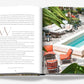 An open book reveals a Bond-like poolside scene from "The Ocean Club" by Assouline, with a woman in an orange dress on the right and text on the left, surrounded by lush plants and elegant outdoor furniture.