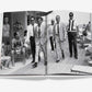 A black and white photo in an open book from Assouline's "The Surf Club" shows men in suits walking on a boardwalk near a beach, surrounded by people in swimwear, capturing the luxury of Miami's iconic club.