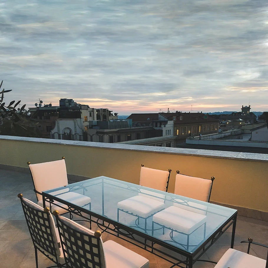 A glass-top table with six Janus et Cie Amalfi Dining Side Chairs in Slate sits on a rooftop terrace, overlooking the city under a cloudy evening sky.
