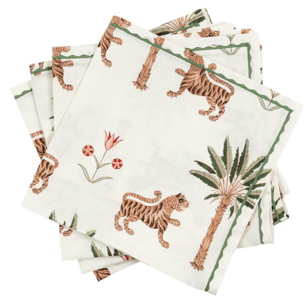 The Matouk Tiger Palm Tabletop Collection features a stack of white napkins with Schumacher designs, showcasing jungle motifs like tigers, palm trees, and green and brown flowers.