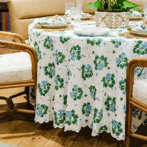A round table with Matouk’s Tillett x Matouk Blue Geranium Table Linen Collection, set with plates and glasses and a potted plant centerpiece, is surrounded by cushioned wooden chairs for an inviting floral look.