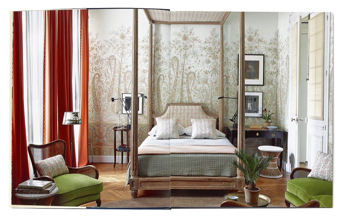 An elegant bedroom featuring a four-poster bed, green armchairs, and floral wallpaper exudes style. Wooden furniture and red curtains add to the decor's warmth. Enhancing the cozy ambiance, a plant and artworks reflect Tom Scheerer's signature blend of relaxed modernism with American interior design elements as illustrated in "The Tom Scheerer Compendium" by Vendome.
