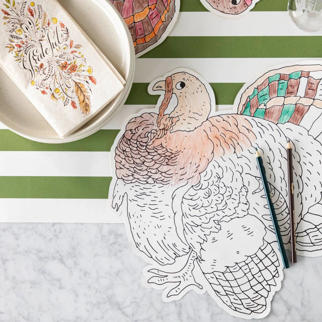 Tabletop featuring the Hester & Cook Die Cut Coloring Turkey Placemat, colored pencils, a striped green and white placemat, a bowl, and a napkin with autumn designs—perfect for a festive holiday crafts coloring activity.