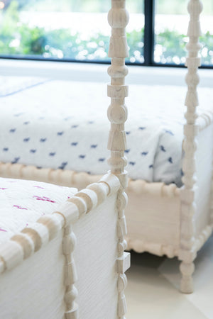 Close-up of the carved corner posts and frames of two St. Tropez Canopy Beds by Mr Brown, featuring patterned white bedding in a bright room.