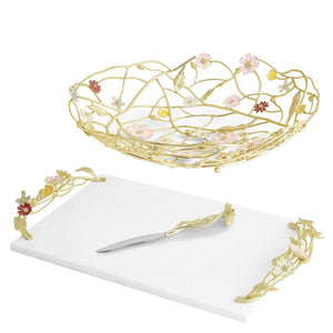 The Michael Aram Wildflower Serveware Collection features a gold wire centerpiece basket with handcrafted wildflowers, an artisanal marble cheeseboard, a knife, and gold floral handles.
