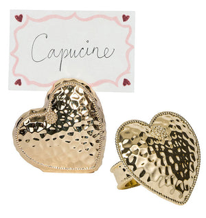 Two Berry & Thread Heart Napkin Rings and Place Card Holders by Juliska, featuring a hammered gold finish and a handwritten "Capucine" card bordered with pink and red hearts.