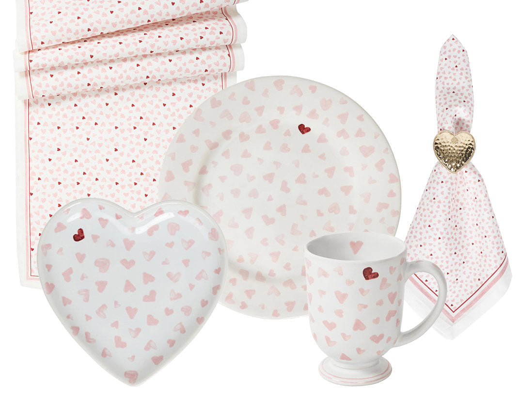 The Juliska Love You More Decor and Tabletop Collection features white plates, a heart-shaped dish, mug, napkin, and table runner adorned with pink and red hearts in a matching sweetheart pattern.