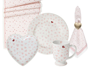 The Juliska Love You More Decor and Tabletop Collection features white plates, a heart-shaped dish, mug, napkin, and table runner adorned with pink and red hearts in a matching sweetheart pattern.