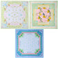 Oh My Mahjong's Mahjong Mat Collection features three square silk scarves: one with butterflies and flowers, one with yellow lemons and leaves, and one inspired by Mahjong mats with a blue border and geometric design.
