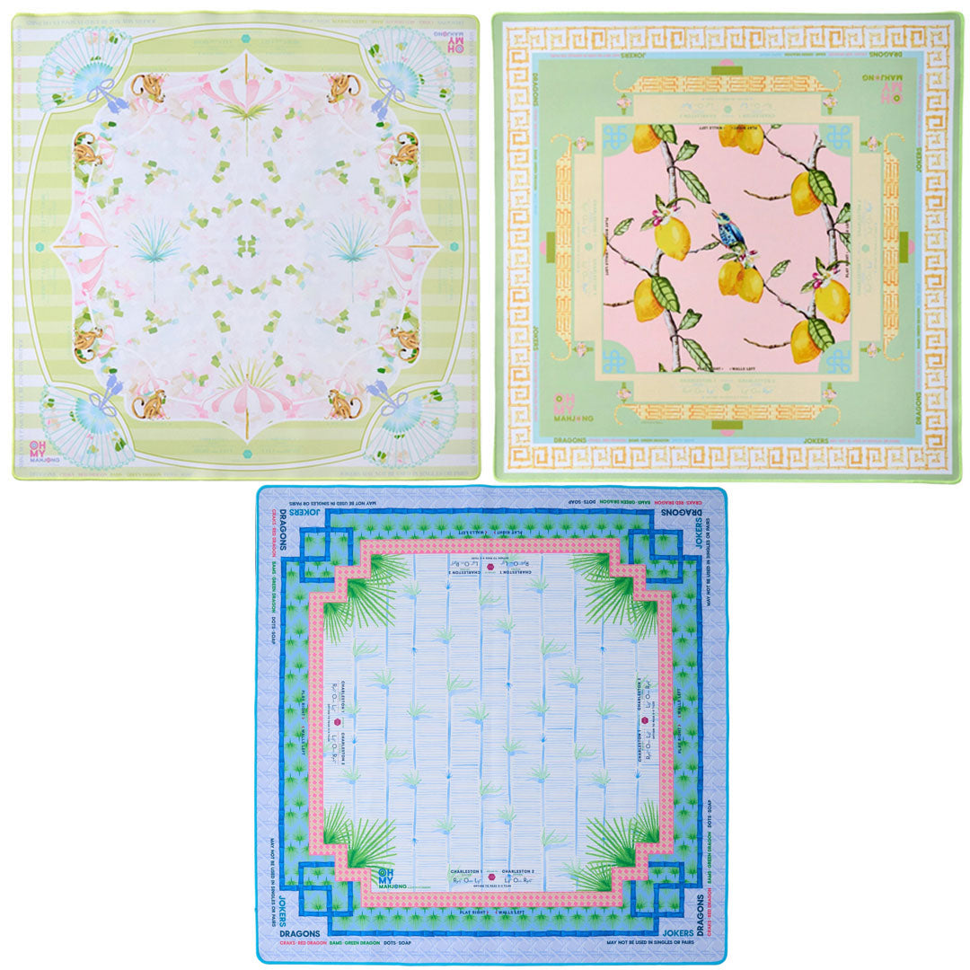 Oh My Mahjong's Mahjong Mat Collection features three square silk scarves: one with butterflies and flowers, one with yellow lemons and leaves, and one inspired by Mahjong mats with a blue border and geometric design.