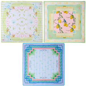 Oh My Mahjong's Mahjong Mat Collection features three square silk scarves: one with butterflies and flowers, one with yellow lemons and leaves, and one inspired by Mahjong mats with a blue border and geometric design.
