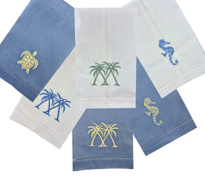 Elevate your table with the Coastal Linen Hand Towel Collection by Pink House Mustique—six blue and white linen towels embroidered with turtles, seahorses, and palm trees for a touch of seaside charm.