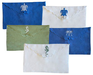 The Linen Envelope Pouch Collection by Pink House Mustique features five clutches in blue, green, and white with embroidered turtles, sea horses, or palm trees. Each secures with a button closure, adding coastal charm to your accessories.