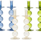 The Kim Seybert Iris Tall Candle Holder Set features five ribbed, hand-blown glass candlestick holders with stacked spheres and scalloped bases in blue, clear, and green, displayed upright against a white background.
