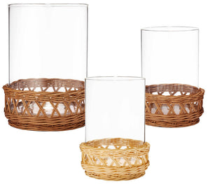 The Navona Rattan Hurricane Collection by Aerin features three clear cylindrical glass vases with decorative rattan pedestal bases in brown tones, blending natural elegance and timeless design against a white background.