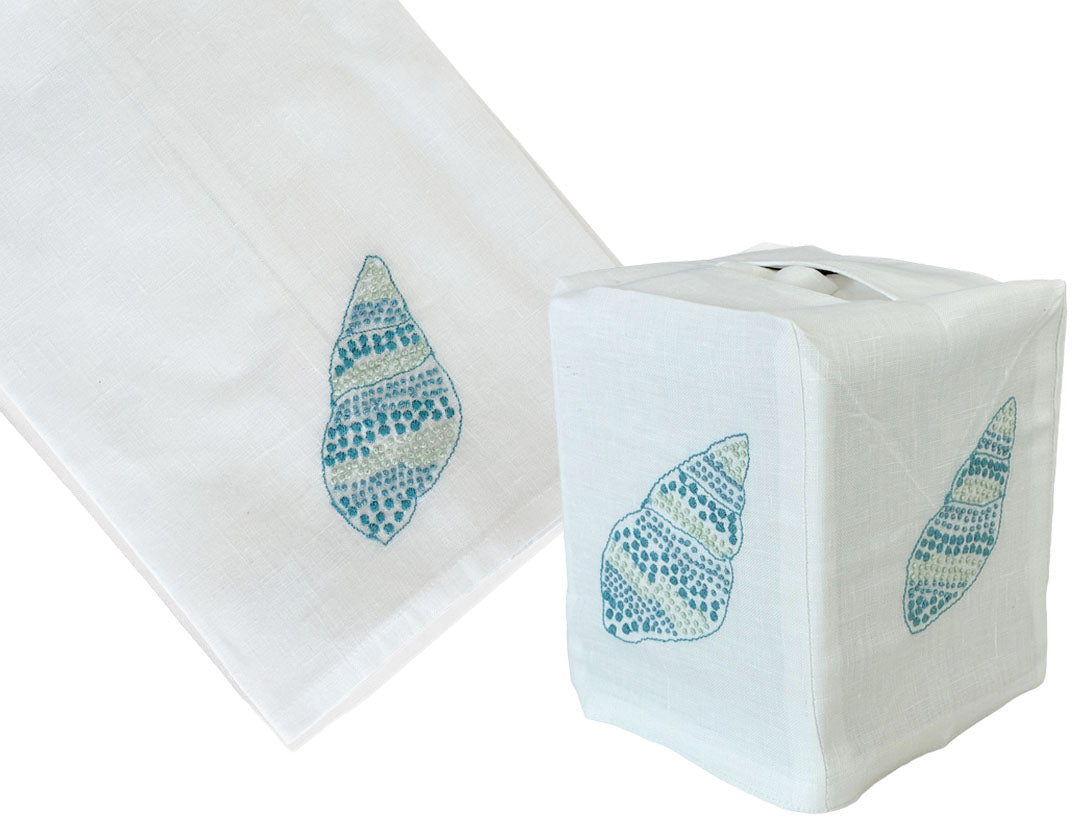 The Haute Home Speckled Conch Bath Collection in Aqua features a white Italian linen tissue box cover and matching towel, both hand embroidered with blue and aqua seashell designs.