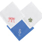 The Dinner Napkin Set by Pink House Mustique includes three folded embroidered napkins: two white linens with pink palm trees and a green turtle, and one blue linen with a white lizard design.