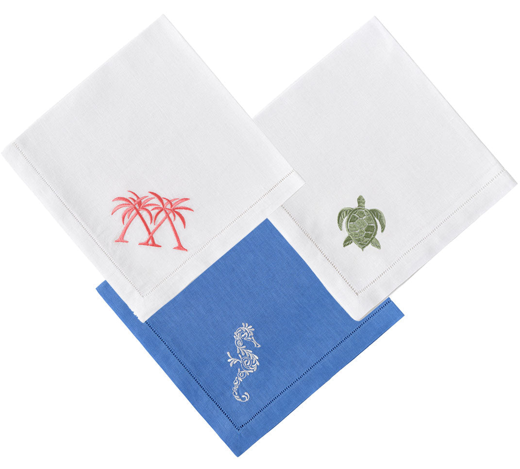 The Dinner Napkin Set by Pink House Mustique includes three folded embroidered napkins: two white linens with pink palm trees and a green turtle, and one blue linen with a white lizard design.