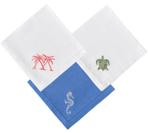 The Dinner Napkin Set by Pink House Mustique includes three folded embroidered napkins: two white linens with pink palm trees and a green turtle, and one blue linen with a white lizard design.