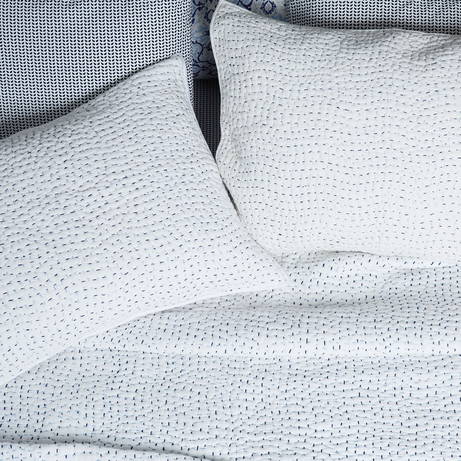 Two white pillows and a matching organic cotton coverlet from the John Robshaw Hand-Stitched Bedding Collection, featuring a subtle stitched pattern, arranged neatly on a textured background.