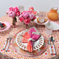 A round table set for four features the Majorelle Tablecloth Collection by Kim Seybert, floral plates, pink-patterned napkins, pink and white glassware, a pink floral centerpiece, and assorted pastries with fruit bowls.