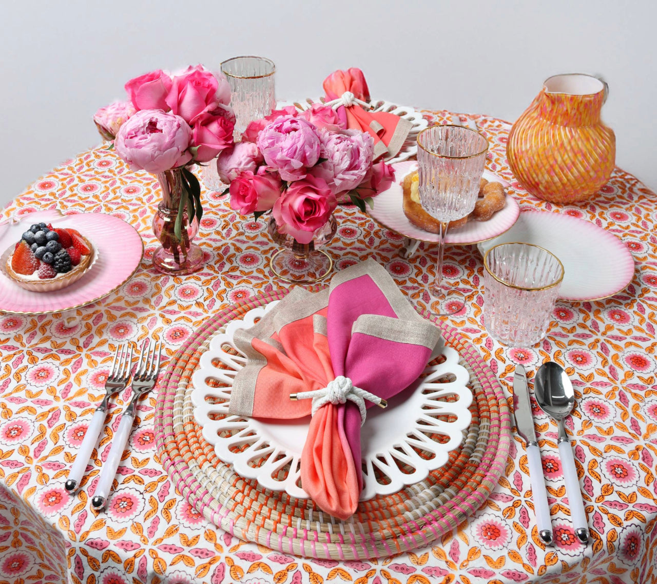 A round table set for four features the Majorelle Tablecloth Collection by Kim Seybert, floral plates, pink-patterned napkins, pink and white glassware, a pink floral centerpiece, and assorted pastries with fruit bowls.
