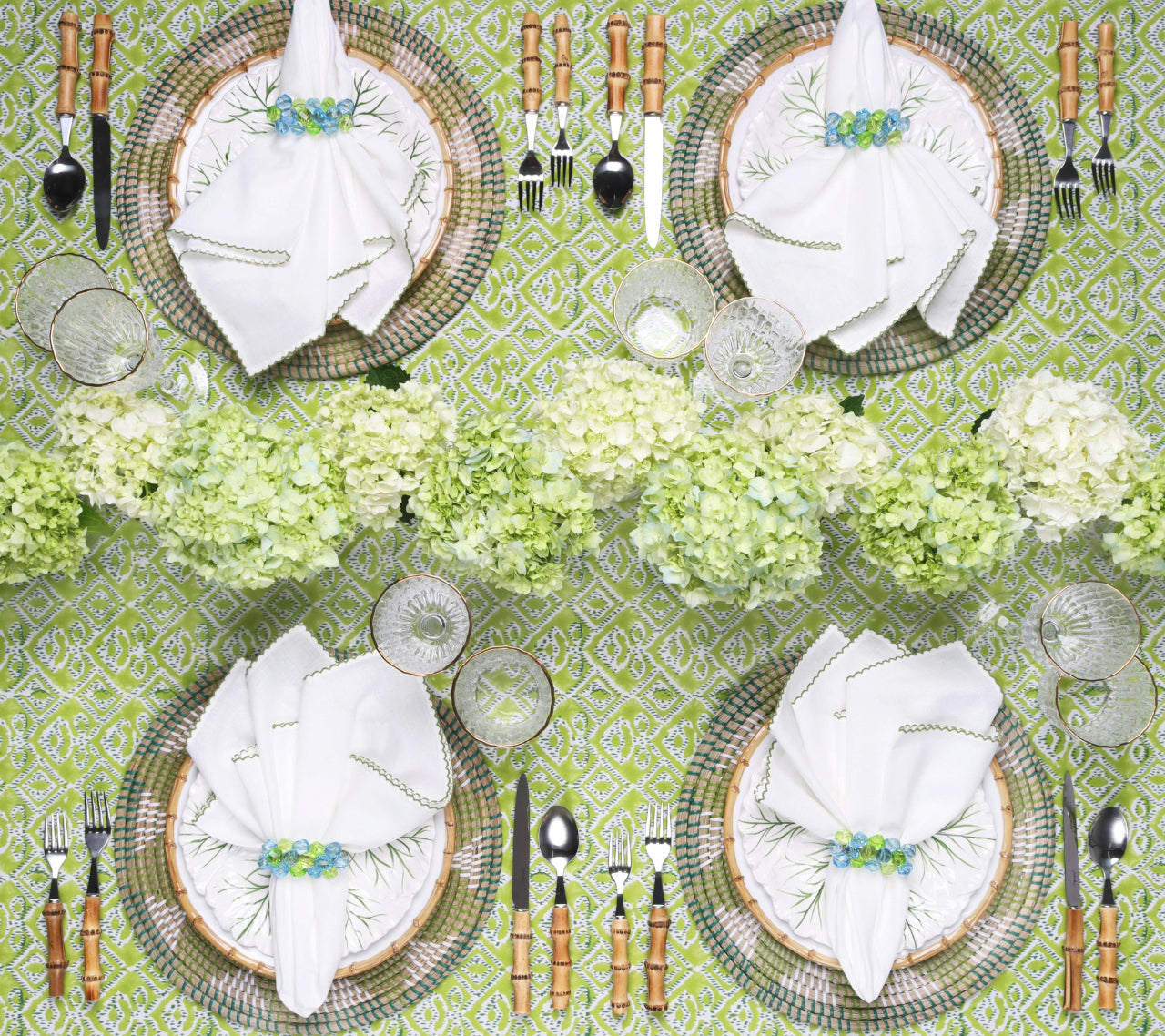 Table set for four with white napkins, woven placemats, bamboo-handled cutlery, clear glassware, and a Kim Seybert Alfresco Tablecloth featuring Indian block printing on 100% cotton, topped with a hydrangea centerpiece.