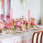 A dining table set with pink flowers, tall pink candles, crystal glassware, and the Kim Seybert Malta Tablecloth Collection, surrounded by wooden chairs in a bright room, creates an elegant table décor.