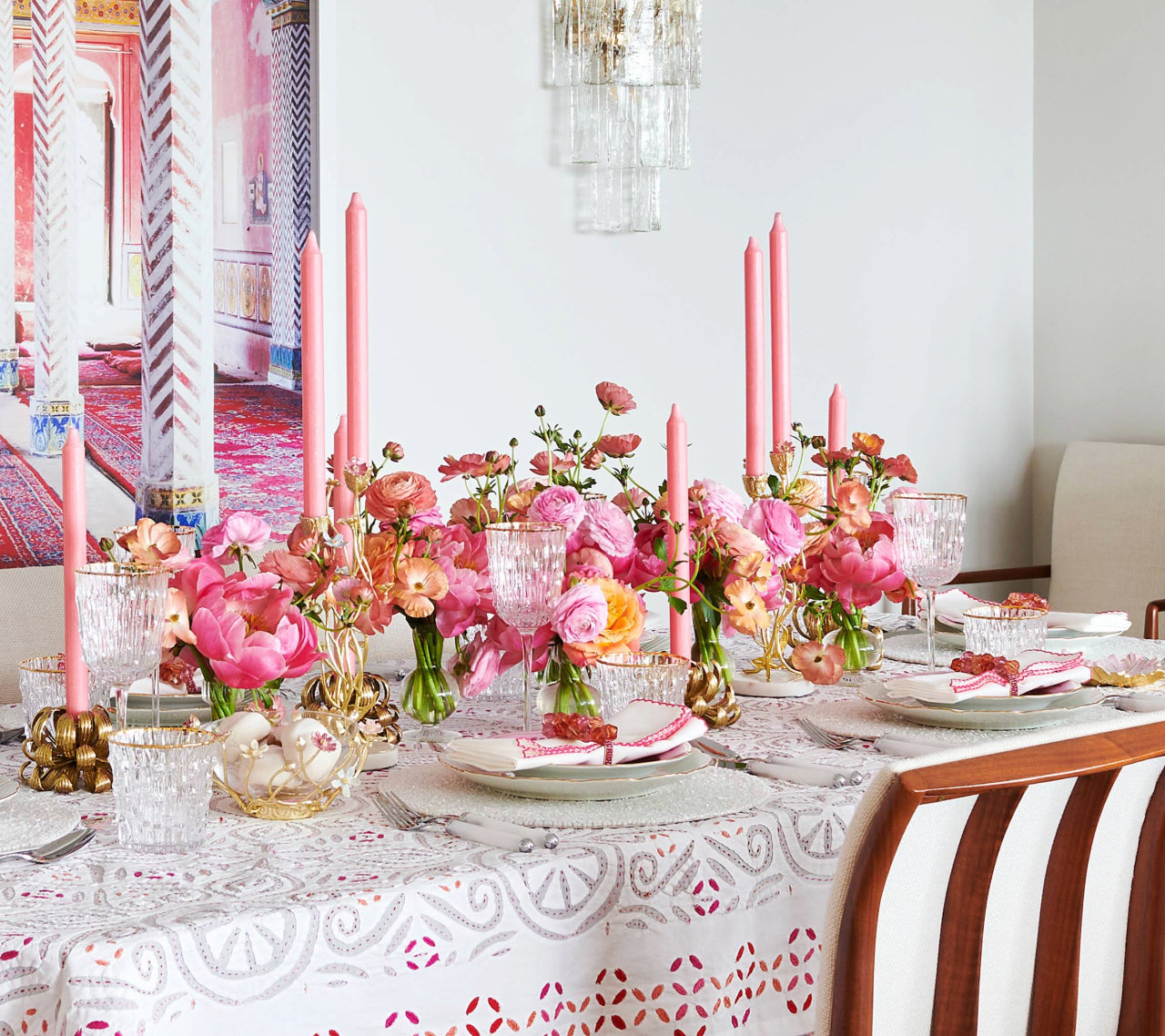 A dining table set with pink flowers, tall pink candles, crystal glassware, and the Kim Seybert Malta Tablecloth Collection, surrounded by wooden chairs in a bright room, creates an elegant table décor.