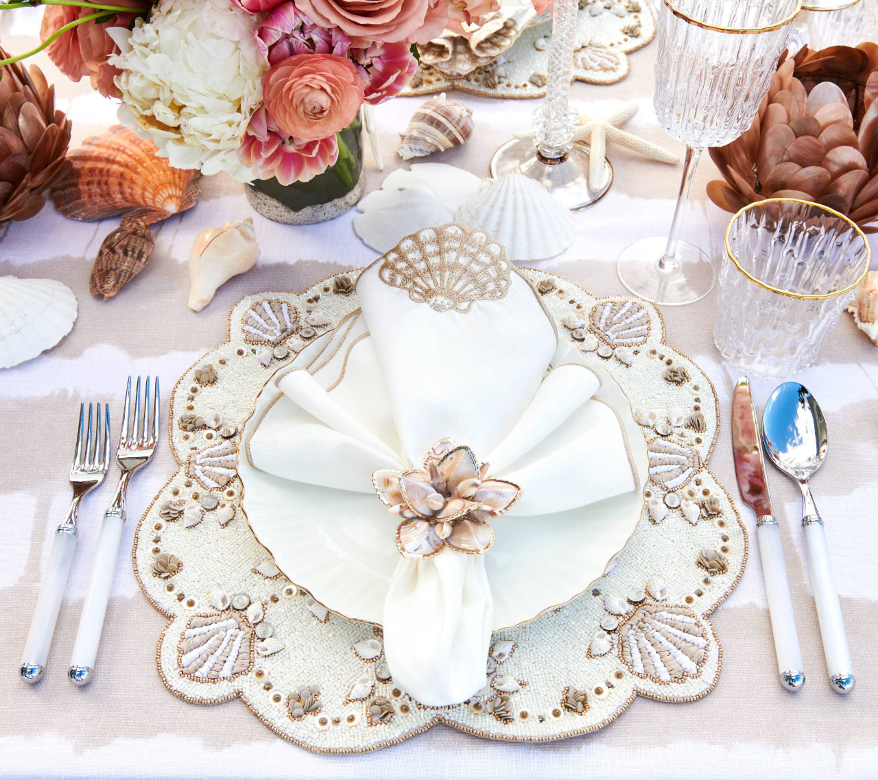 A formal table setting with a Kim Seybert Beachcomber Placemat, white napkin and ring, floral centerpiece, glassware, and silverware on a striped tablecloth—ideal for elevating any coastal home.