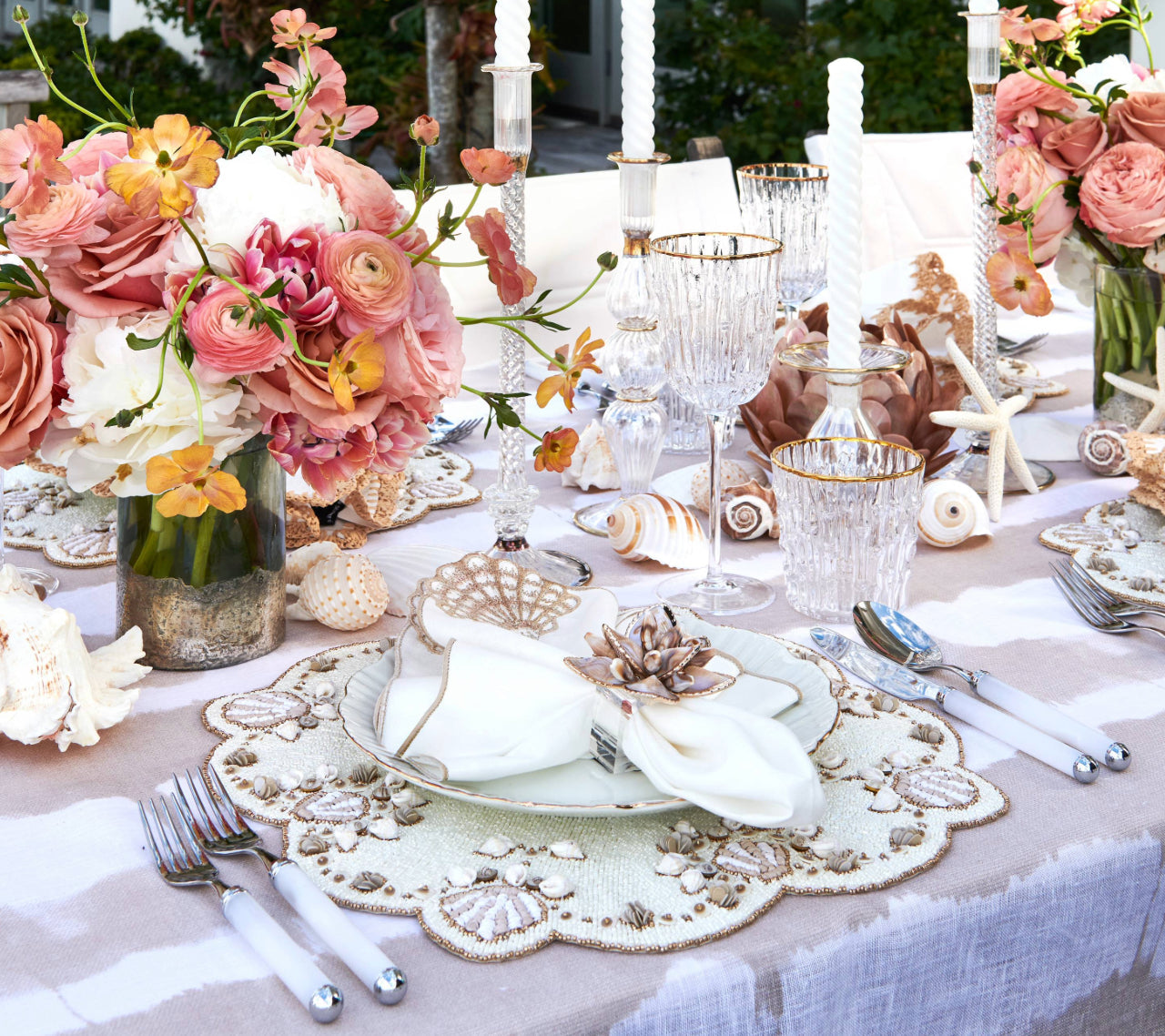 A formal table is set with floral centerpieces, crystal glassware, seashell decor, candles on a beige tablecloth, and white napkins accented by the Kim Seybert Beachcomber Napkin Ring Set.