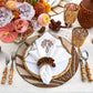 A table setting features a white plate, patterned placemat, bamboo-handled cutlery, embroidered napkin with the Kim Seybert Royal Palm Napkin Ring, floral centerpiece, and amber glassware for chic tropical style.