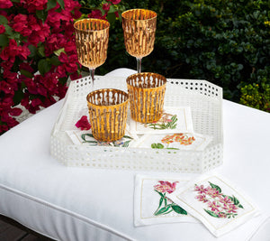 A white tray displays three Kim Seybert Gold Bamboo Tumblers with hand-applied 24k gold and four embroidered floral napkins on a white cushion, set against pink flowers and greenery.