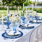 An outdoor dining table features blue and white plates, cobalt blue beads, blue glassware, white flowers, tall white candles, clear chairs, and the Kim Seybert Lagoon Placemat for an ocean-inspired touch amid lush greenery and water views.