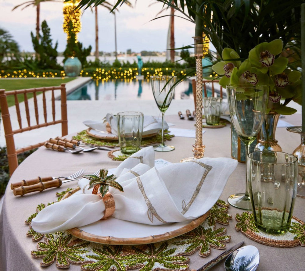 Elegant outdoor dining set with nature-inspired decor, green glassware, white napkins featuring the Kim Seybert Palm Coast Napkin Ring (Set of 4), and a pool with string lights in the background.