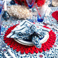 A table set with Kim Seybert Majorelle Napkin Set in blue and white, red placemats, blue glassware, silver cutlery on a floral tablecloth, with coral and flowers as centerpieces.
