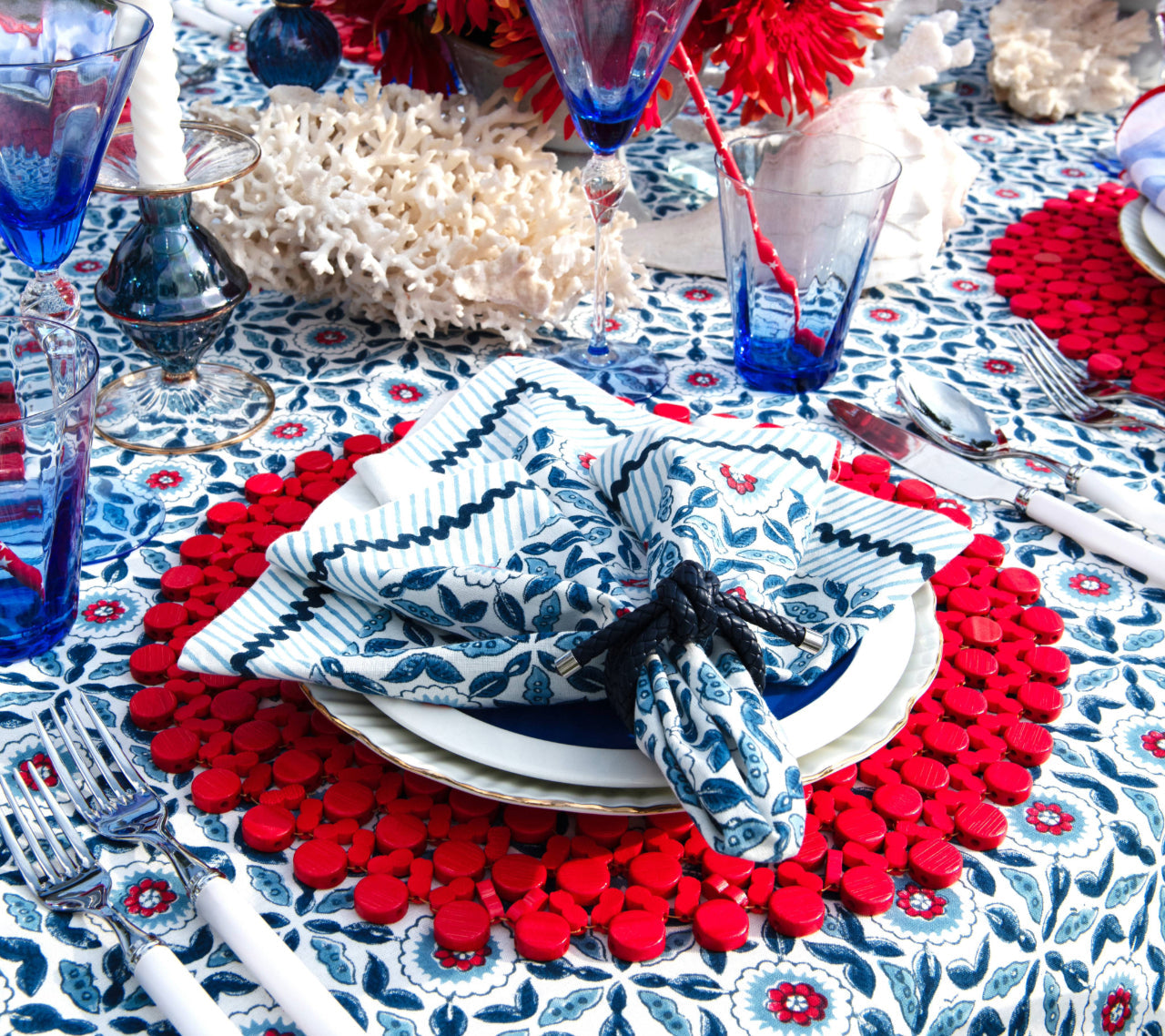 A table set with Kim Seybert Majorelle Napkin Set in blue and white, red placemats, blue glassware, silver cutlery on a floral tablecloth, with coral and flowers as centerpieces.