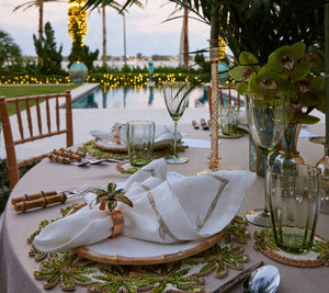 A sophisticated outdoor dining table is beautifully arranged with floral decorations, green glassware, and folded napkins on Kim Seybert's Oasis Placemats by the poolside, framed by palm trees and illuminated string lights in the background, crafting an ideal setting for tropical entertaining.