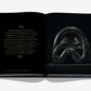 Assouline's 'Uzbekistan: Masterpieces of the Silk Road' presents an open book with black pages that captures a quote about the historical symbolism of fertility and birth. On the right page, a metallic sculpture featuring two snake-like figures is showcased, echoing the rich tapestry of discoveries along the ancient Silk Road and reminiscent of archaeological finds in Uzbekistan.