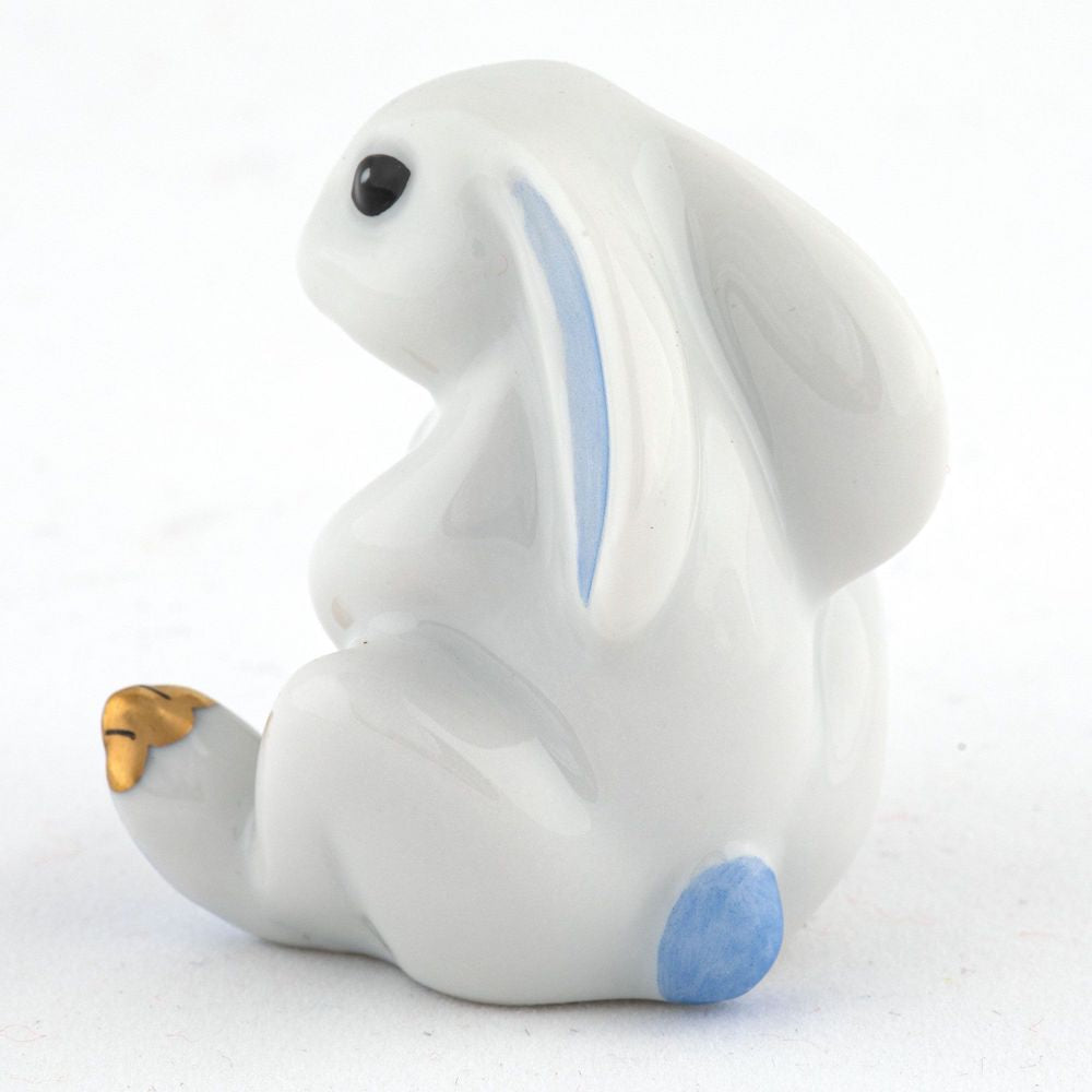 The Herend Bunny with Cross figurine, sitting upright, exemplifies artistic craftsmanship with its hand-painted designs. Blue accents decorate the ears and tail, and a gold front paw adds elegance.