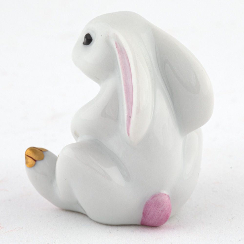 The Herend Bunny with Cross is a sitting ceramic figurine featuring hand-painted pink and gold accents on its ears and feet, reminiscent of Herend porcelain artistry, against a plain white background.