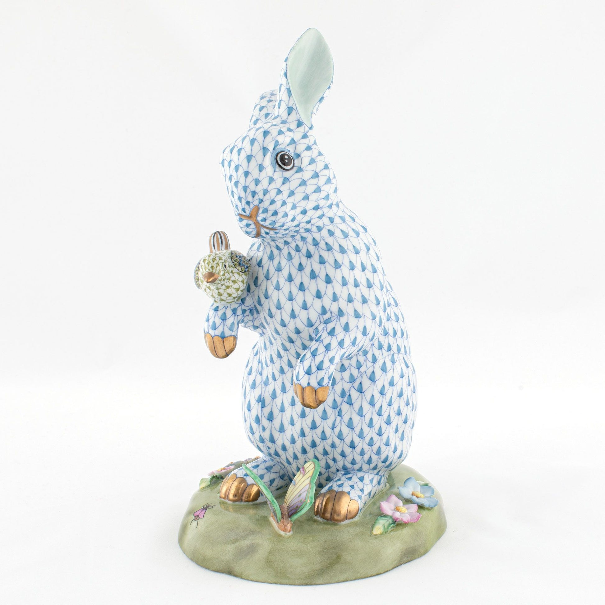 The Herend Reserve Collection Anniversary Bunny is a hand-painted porcelain figurine depicting a standing rabbit with blue and white patterns, holding a decorated egg on a grassy, flowered base. Inspired by classic Herend designs.