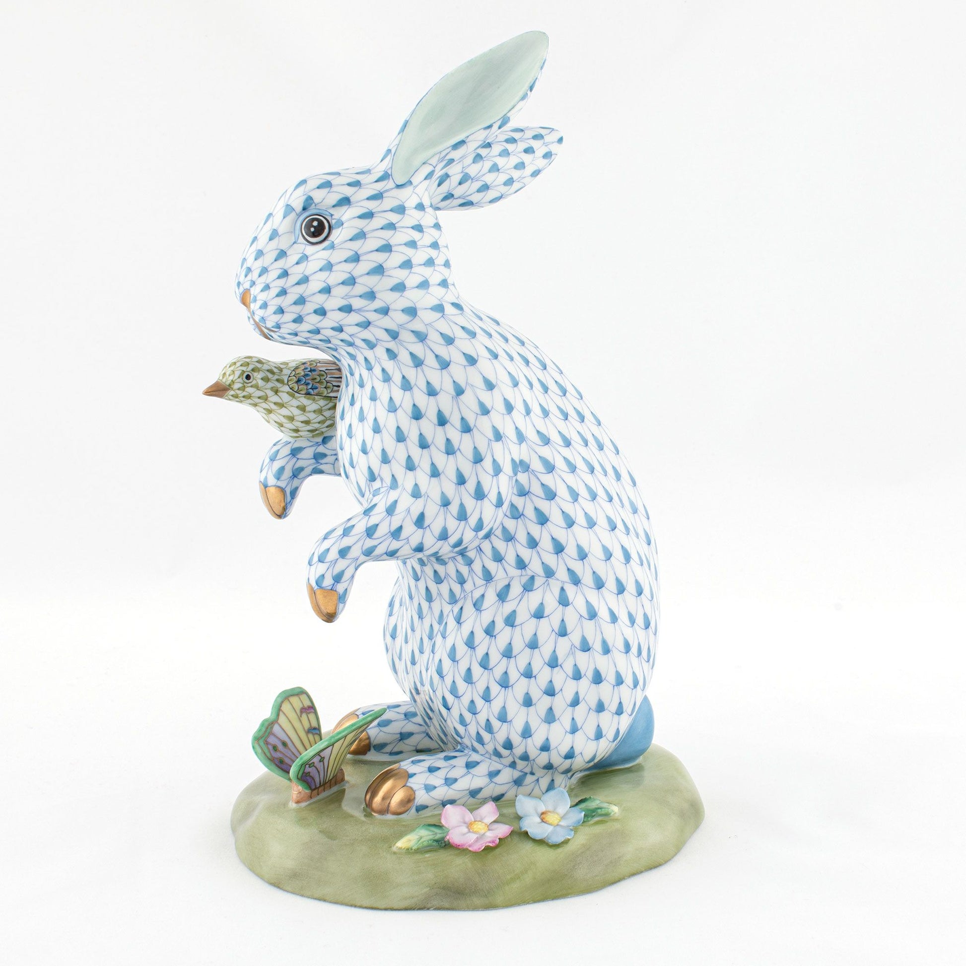 The Herend Reserve Collection Anniversary Bunny features a blue and white patterned rabbit with a bird on its paw, standing on a floral base with leaves and butterfly accents, showcasing exquisite hand-painted designs.