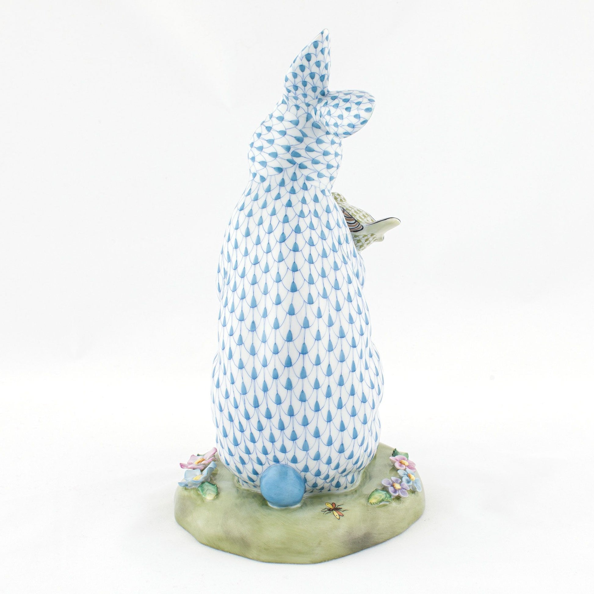 The Herend Reserve Collection Anniversary Bunny features blue and white hand-painted scales, sits upright on a grassy base decorated with flowers and an insect, and is shown from the back.