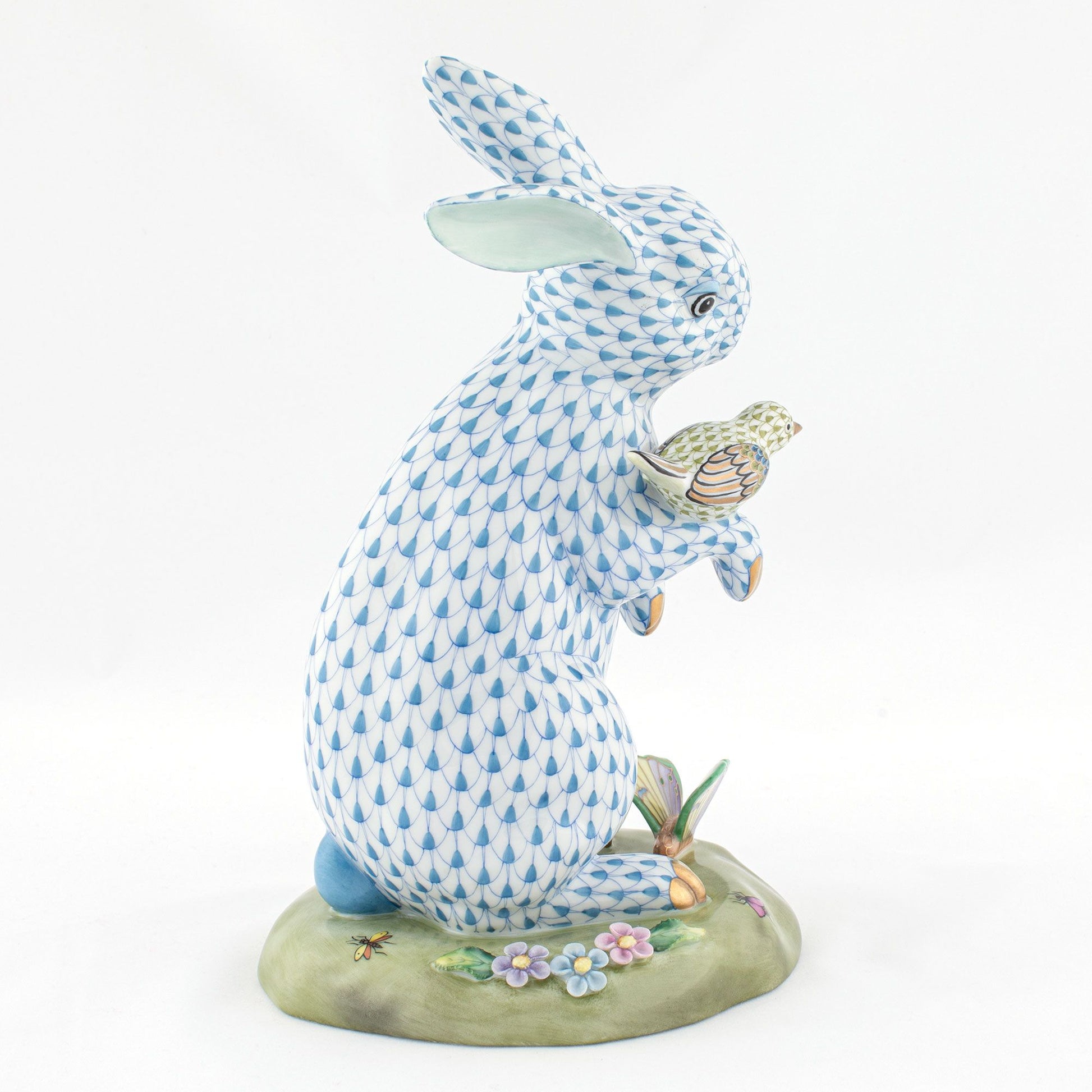 The Herend Reserve Collection Anniversary Bunny is a blue and white porcelain rabbit with a small bird, standing on a floral green base with a butterfly, featuring exquisite hand-painted designs inspired by classic Herend porcelain.