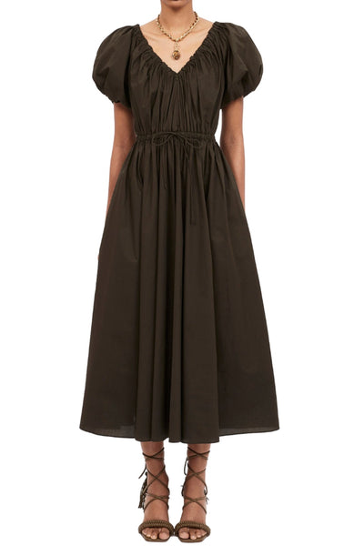 Person wearing the Ulla Johnson Vika Militaire Dress featuring a dark green hue with puff sleeves, a V-neck, and a gathered waist, complemented by lace-up sandals.