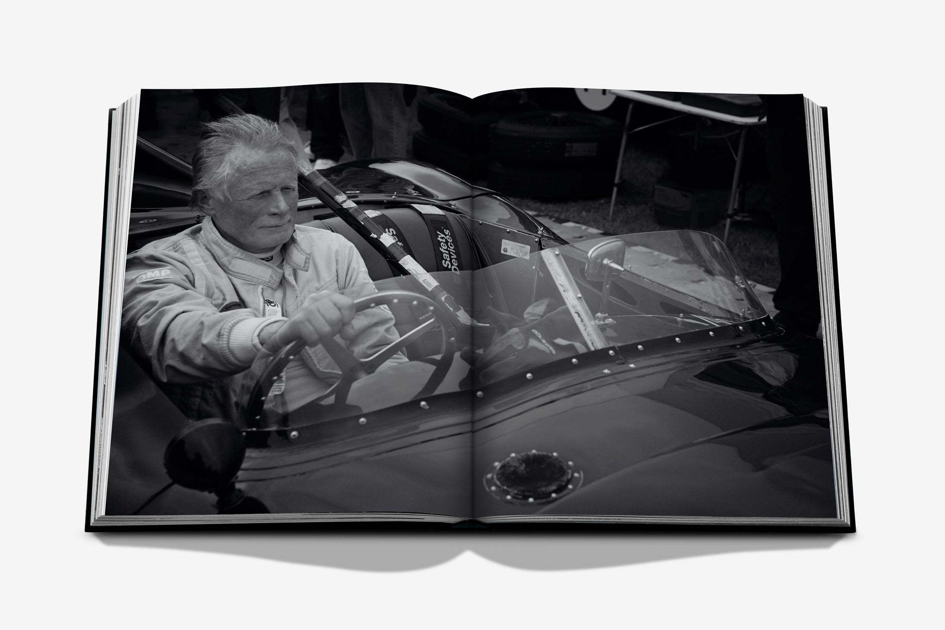 A black and white photograph of an older man in a racing suit seated in a vintage race car is featured on the open pages of Vintage Cars (2nd Edition) by Assouline, set against a white background—a timeless image that embodies the essence of automotive history.