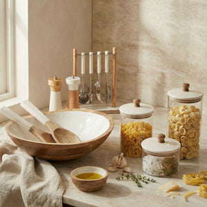 The Be Home Madras Bar Tool Set is displayed on a marble countertop, alongside wooden and ceramic kitchenware, glass jars of pasta and spices, olive oil, and utensils in a sunlit kitchen.